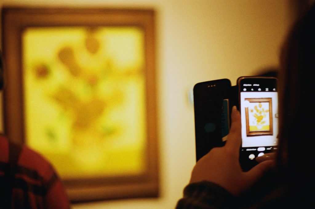 Vincent Vag Gogh's painting Sunflowers in a 2019 Tate Britain exhibition. The painting is out of focus. In the foreground a visitor to the gallery takes a photograph with a mobile phone