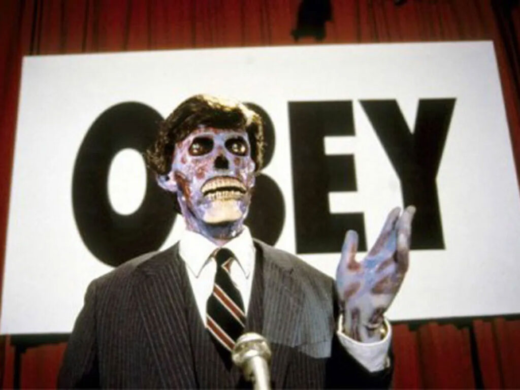 Still from John Carpenter's film They Live. A businessaman in a pin-stripe suit and waistcoat standing at a microphone is revealed to be one of the invading aliens repressing and killing humanity. Behind him a sign reads 'OBEY'