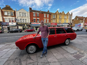 A man proudly leans on his beautifully-restored 1970s Hillman Hunter estate car, in a deep orange colour. Behind him the A5 road in North London and a row of shops.