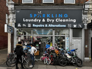 Dry cleaner's shop in London with many bikes stacked up for sale outside on the pavement. Two kids are in front of the shop